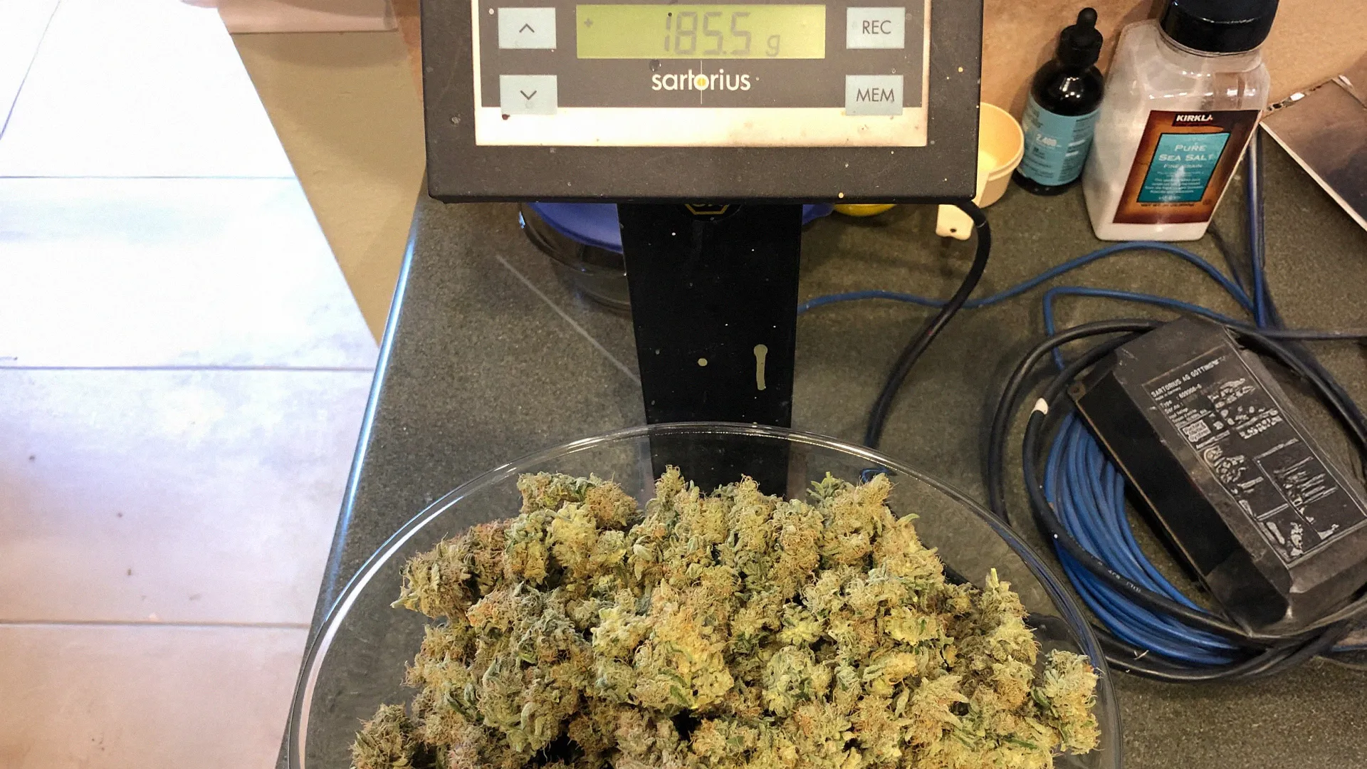 A digital scale displaying 185.5 grams with a bowl of cannabis buds on it, surrounded by various items on a countertop.