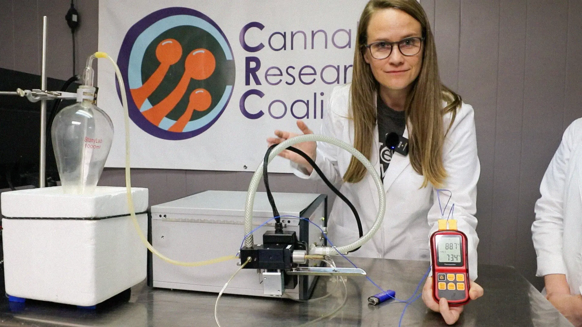 A scientist in a lab coat demonstrates equipment, holding a digital device, with a "Cannabis Research Coalition" banner in the background.