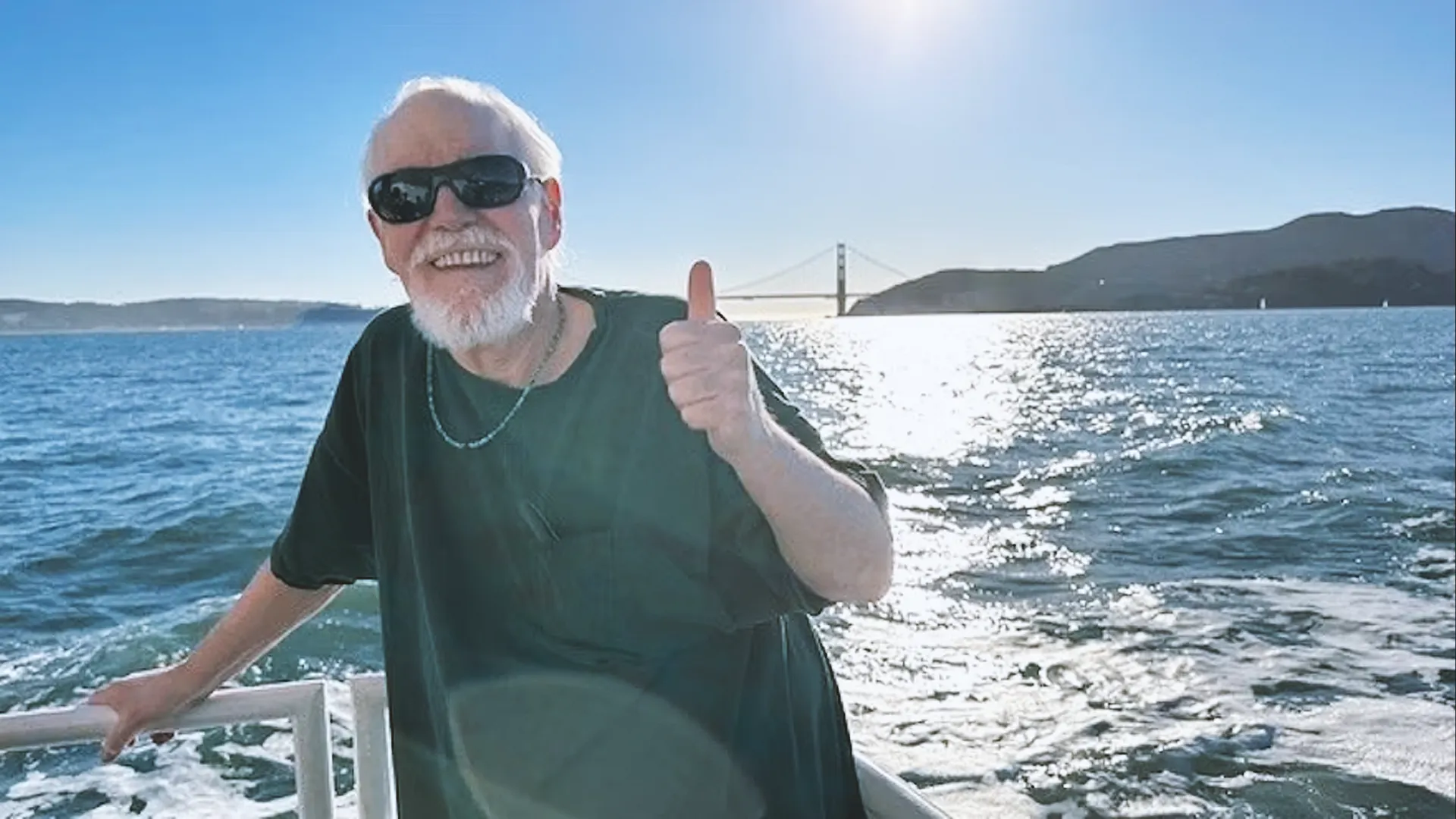 Elderly man with sunglasses gives a thumbs-up on a boat with the Golden Gate Bridge and a sunny sky in the background.
