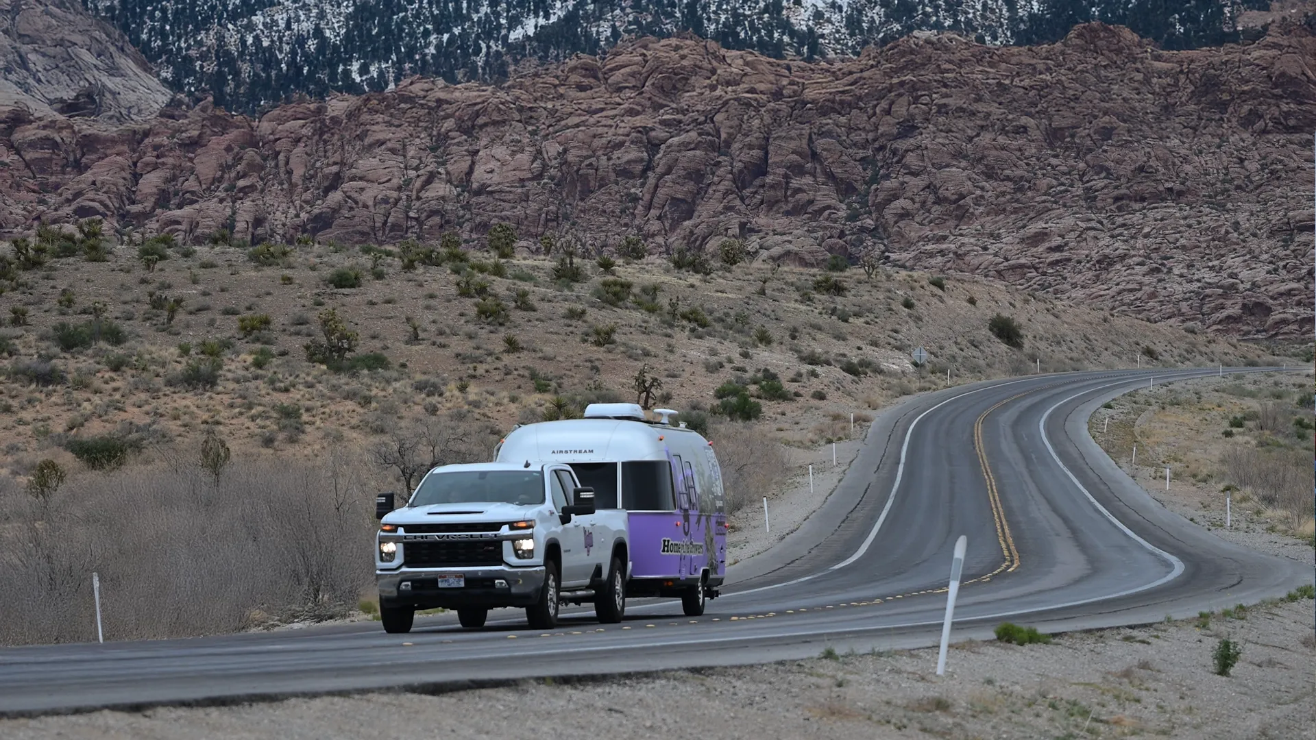 A white truck towing a purple trailer on a winding desert road with rocky hills and sparse vegetation in the background.