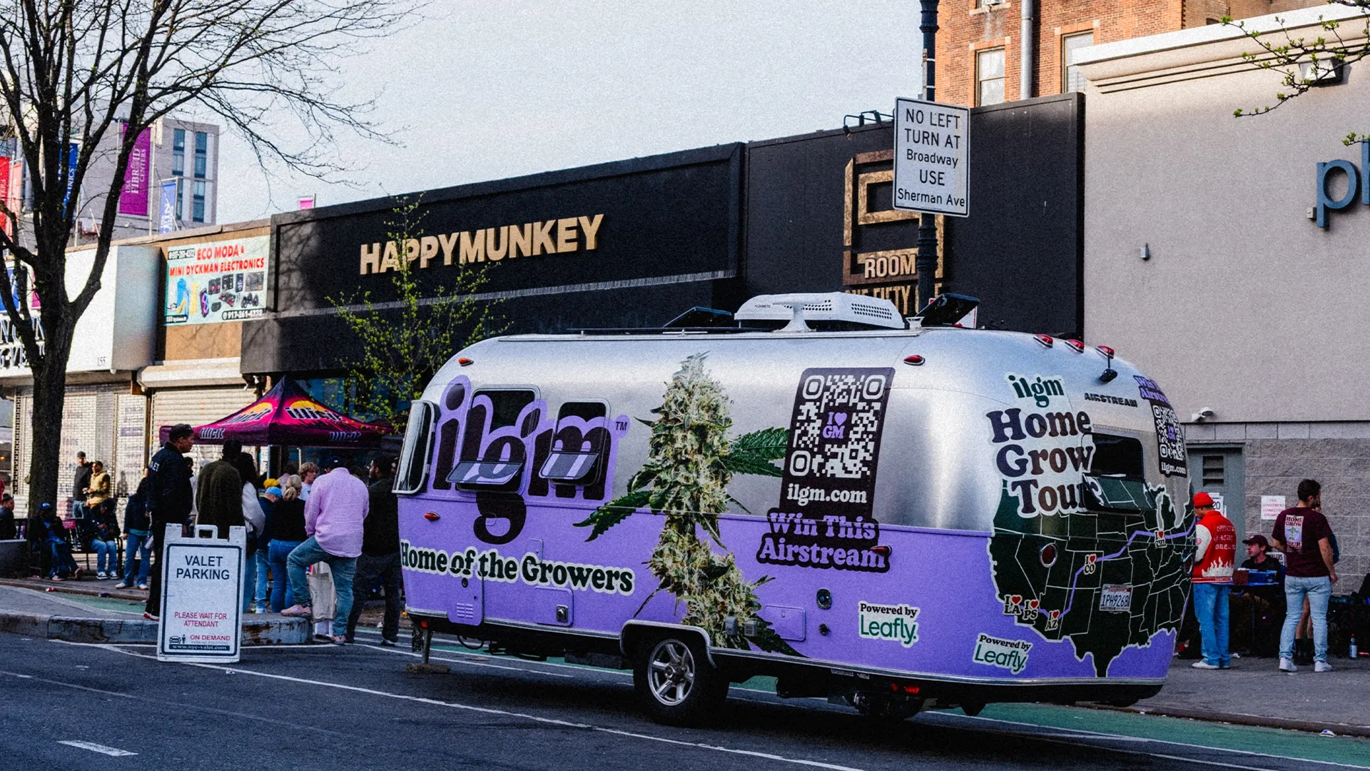 A purple trailer with "Home Grow Tour" branding is parked on a street near a valet parking sign, with people and buildings in the background.