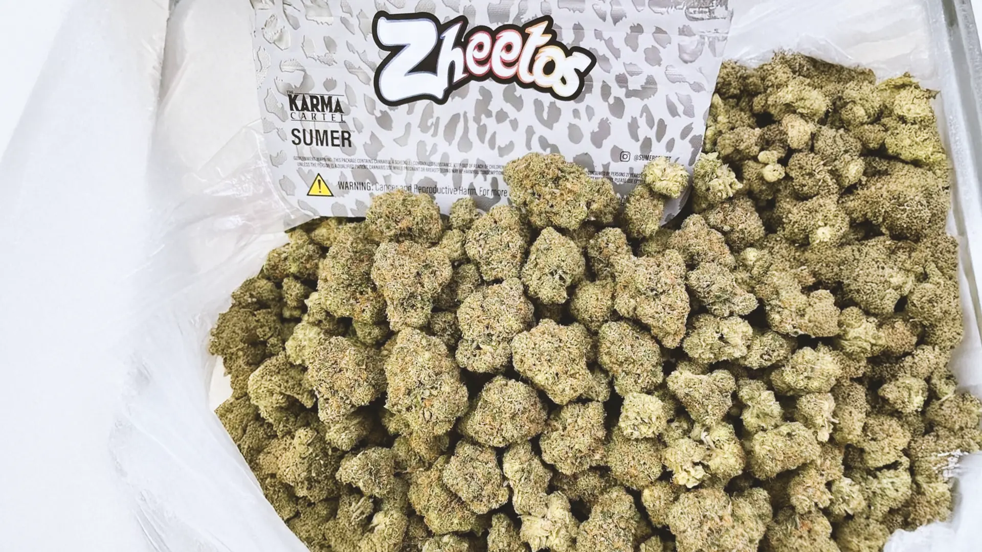 A large pile of cannabis buds next to a Zheetos branded package with leopard print design, labeled "Karma Genetics Sumer."