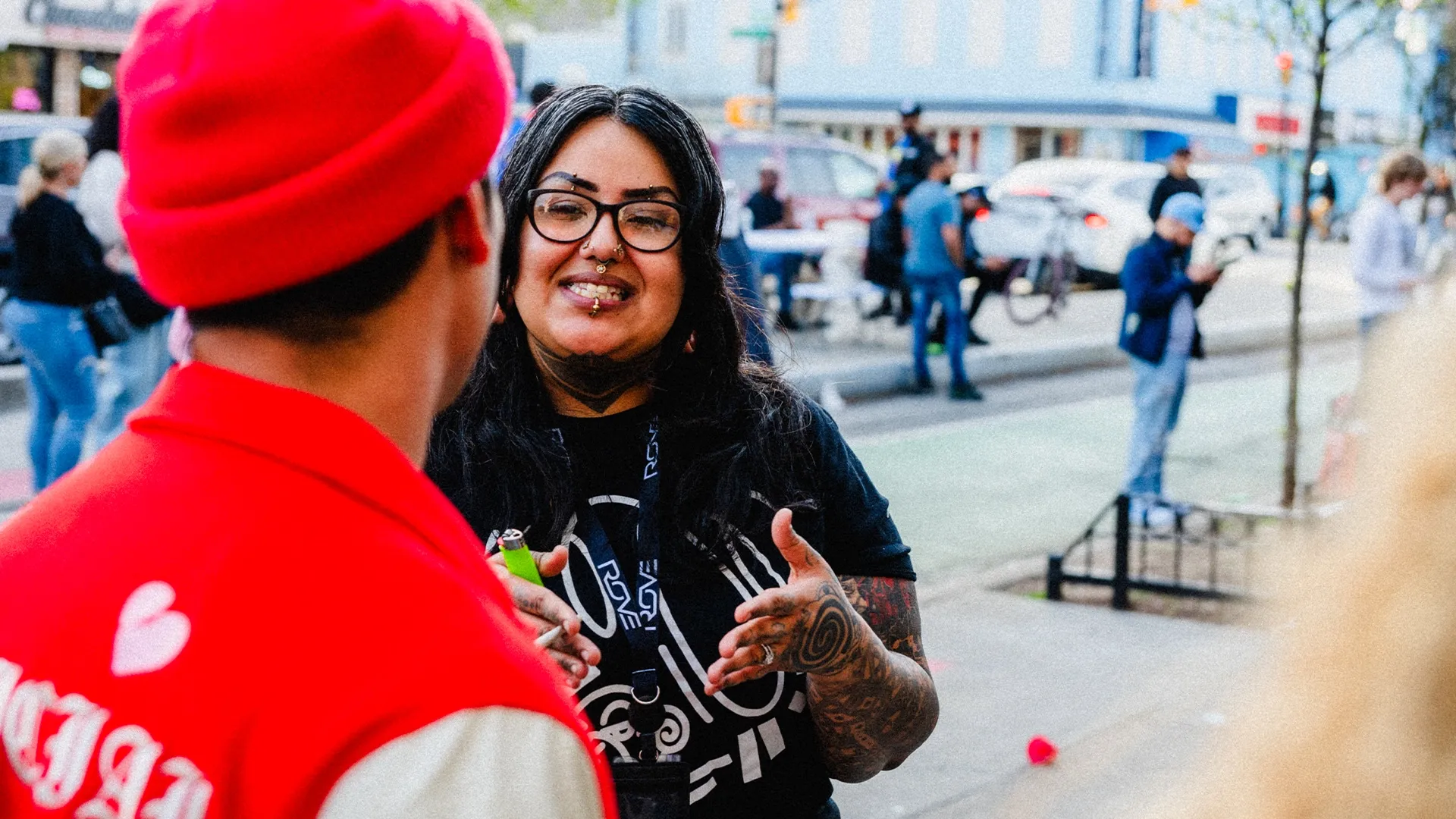 A woman with glasses and tattoos talks to a person in a red jacket on a city street, with people and buildings in the background.