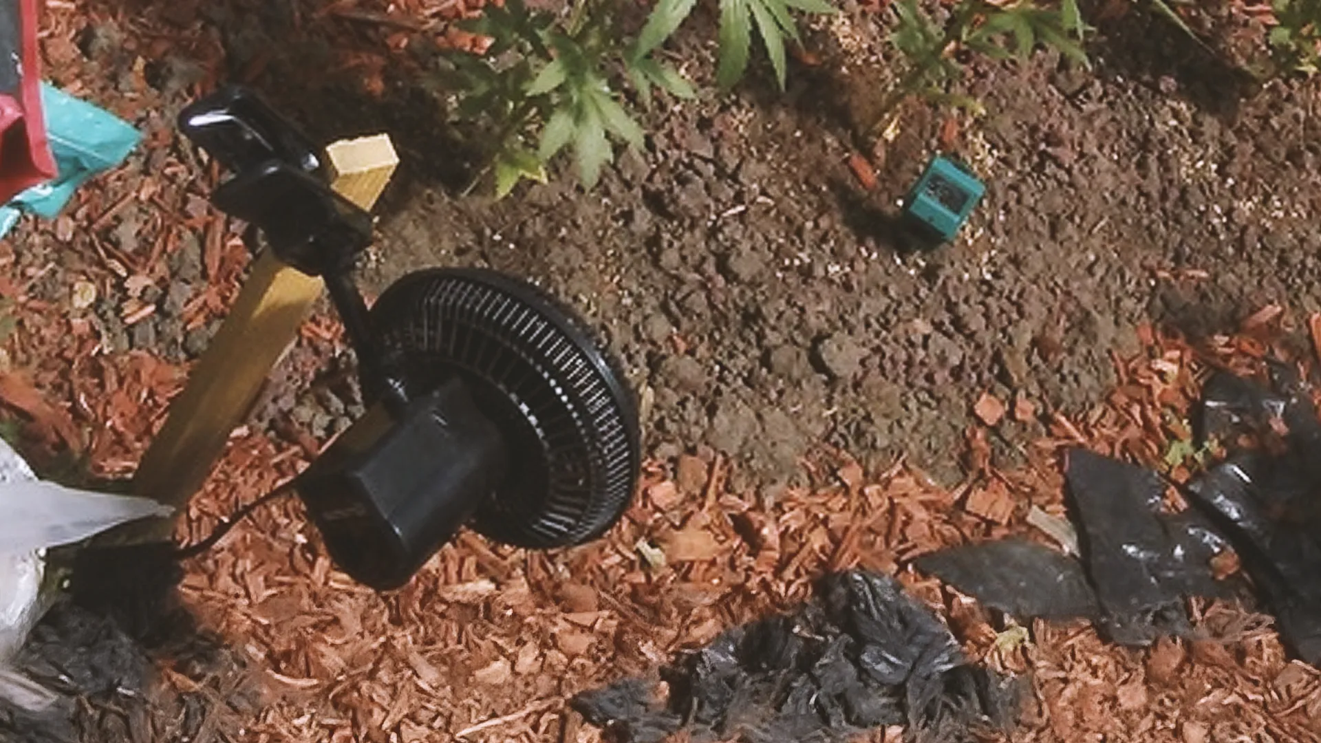 A black fan attached to a wooden post in a garden with soil, mulch, and a small green plant in the background.