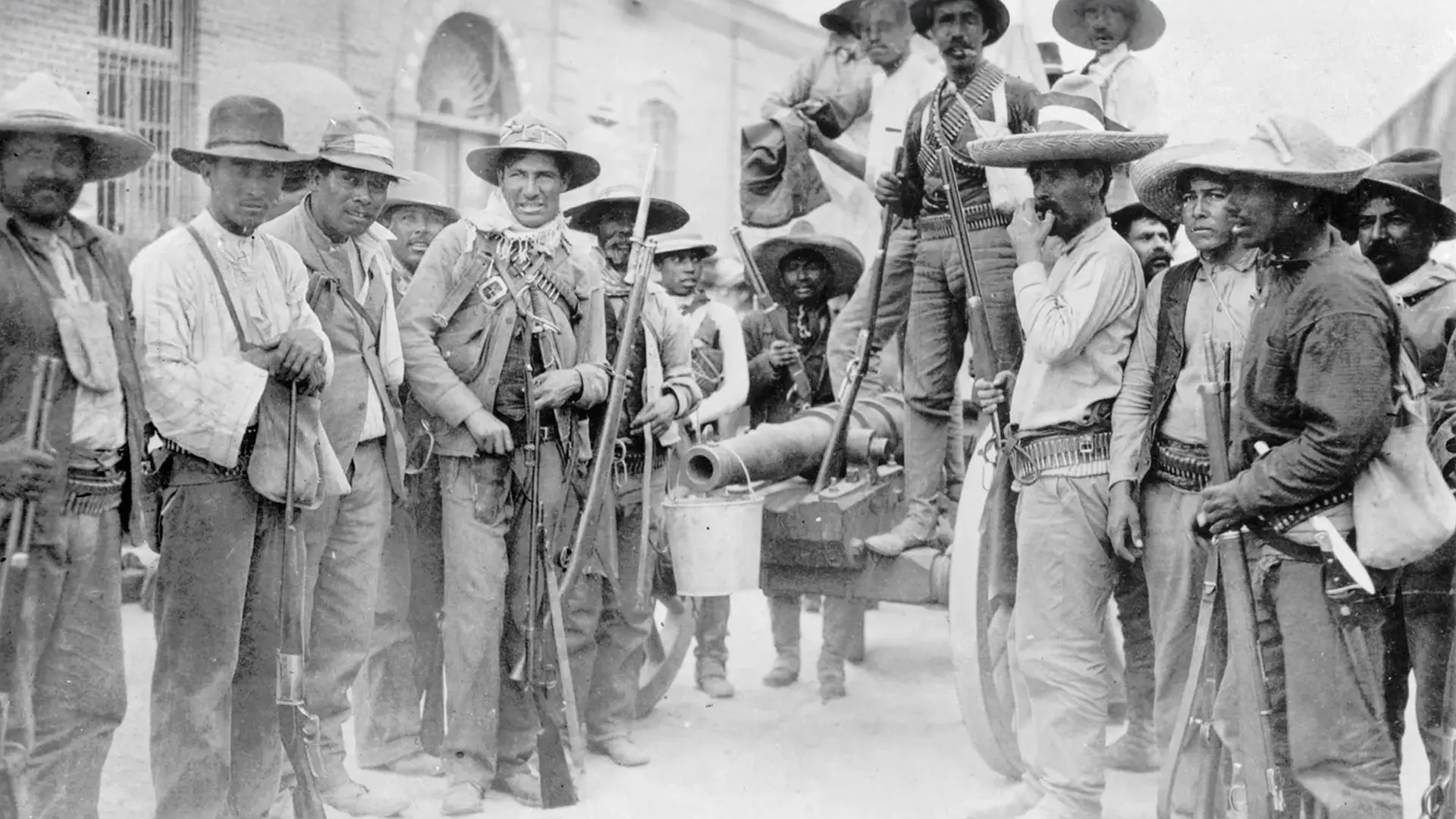 A group of armed men in traditional attire pose with rifles and a cannon in a historical black-and-white photo.