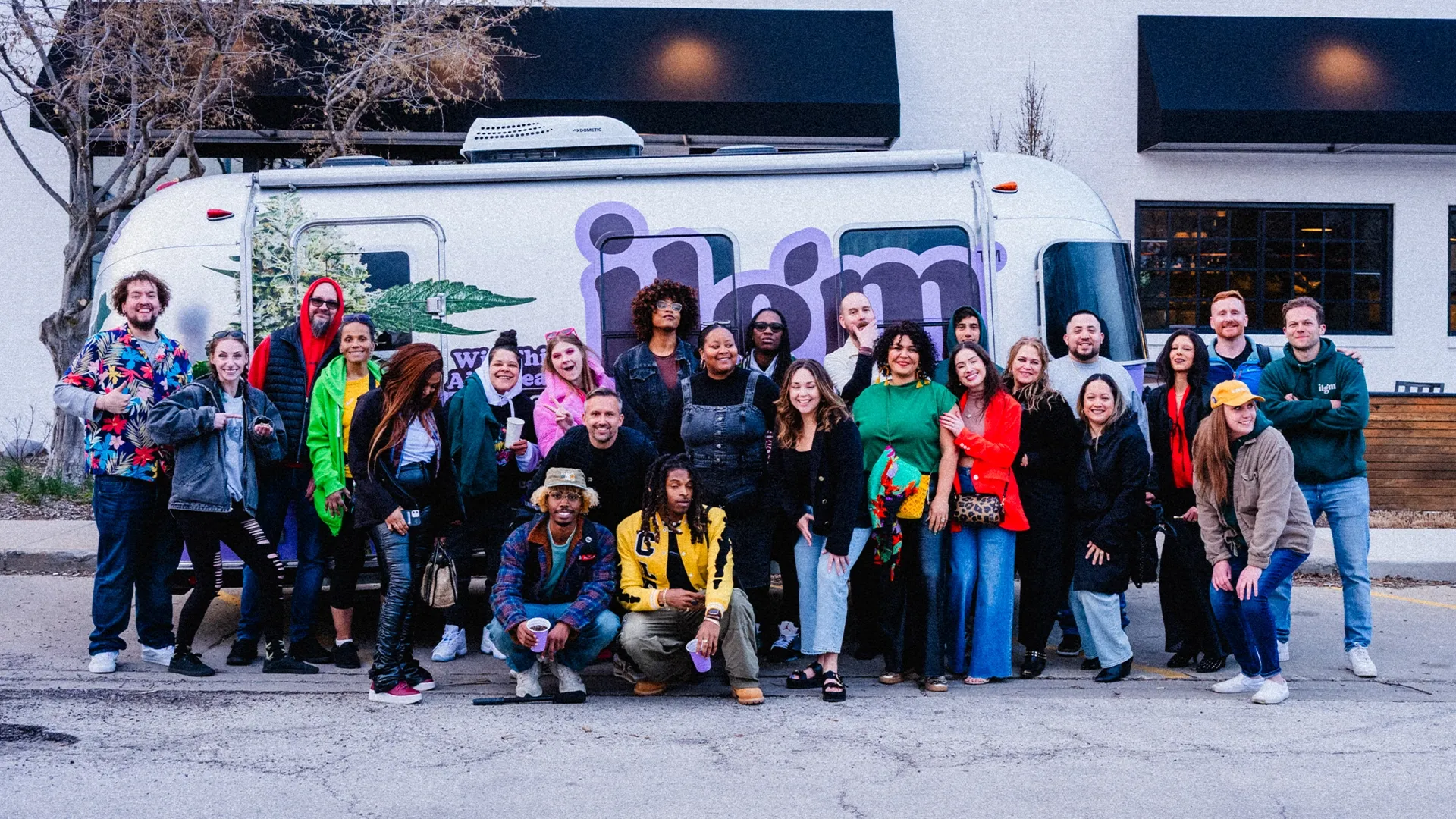 A diverse group of people posing cheerfully in front of a white trailer with colorful graffiti. Some are crouching, others standing.