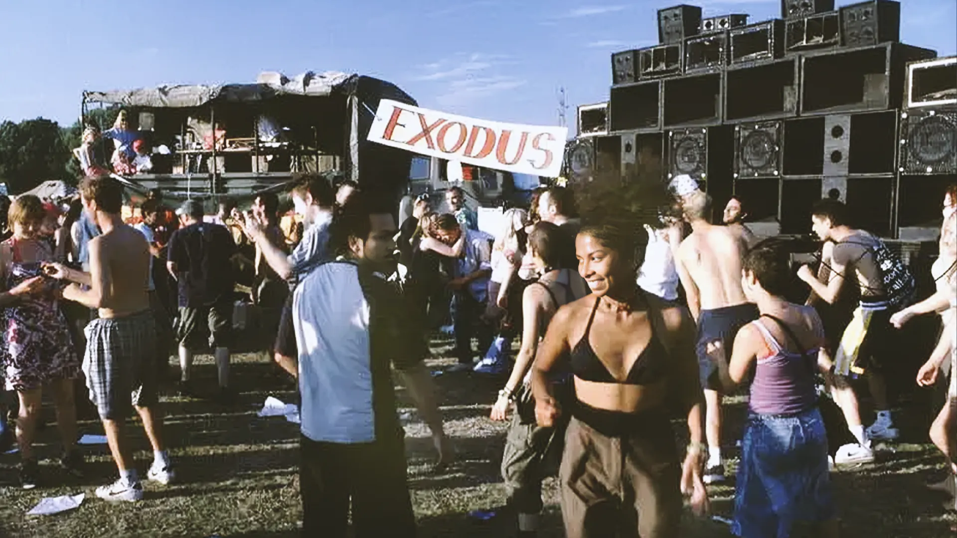 Outdoor rave with people dancing near large speakers and a banner reading "Exodus." The scene is lively under a clear blue sky.