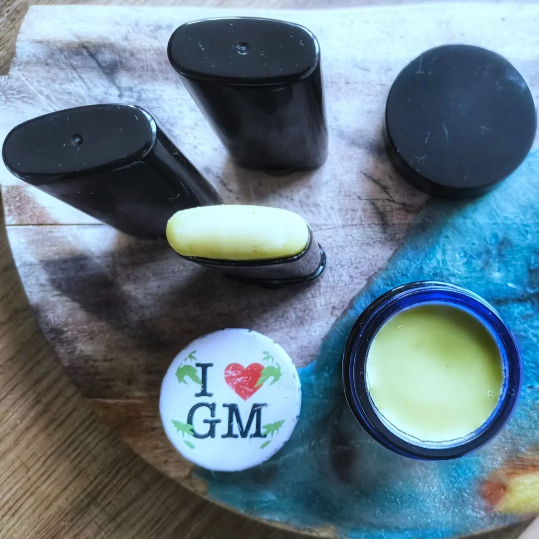 Overhead shot of black deodorant sticks, an exposed yellow stick, an open blue jar of balm, and a white "I ♥ GM" badge on a wooden board.