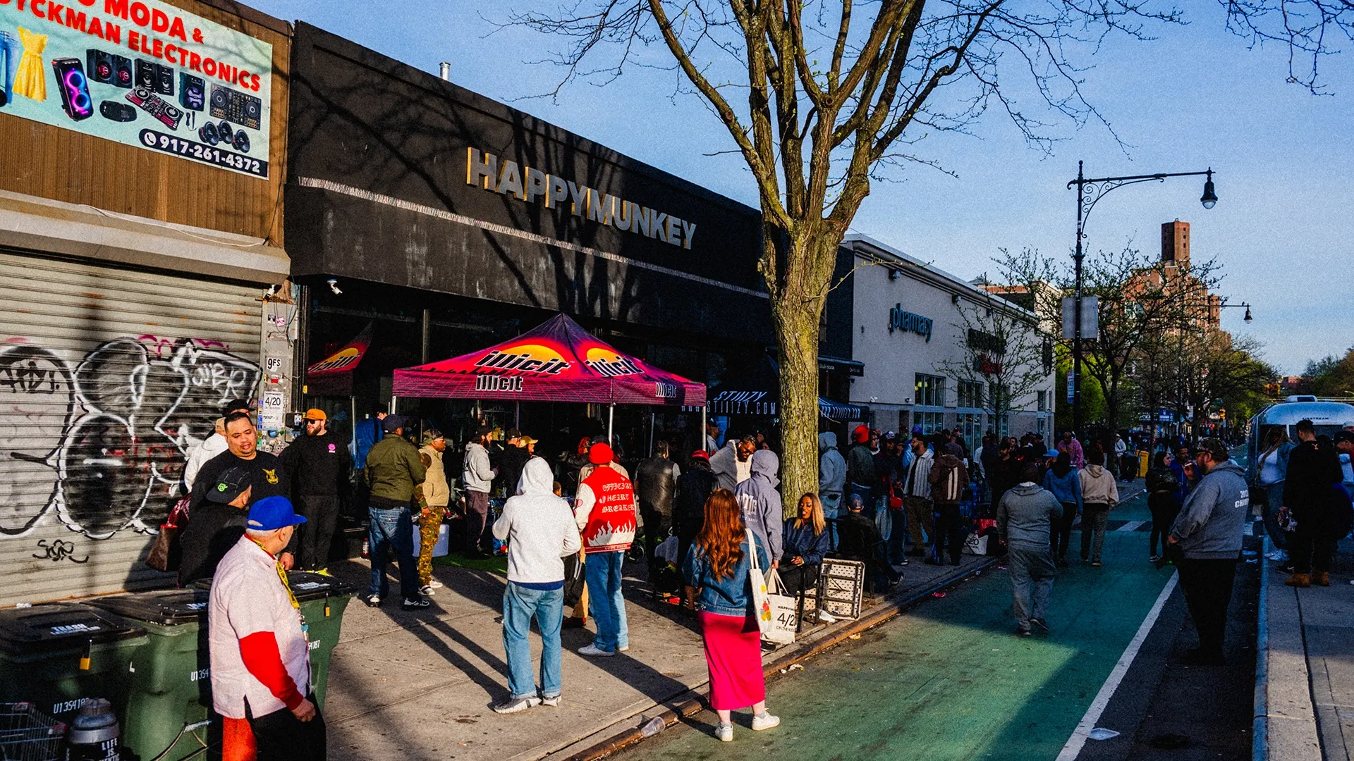 People gather outside a store with a "Happy Munkey" sign, under a Juicy tent on a sunny day. Graffiti is visible on a nearby shutter.