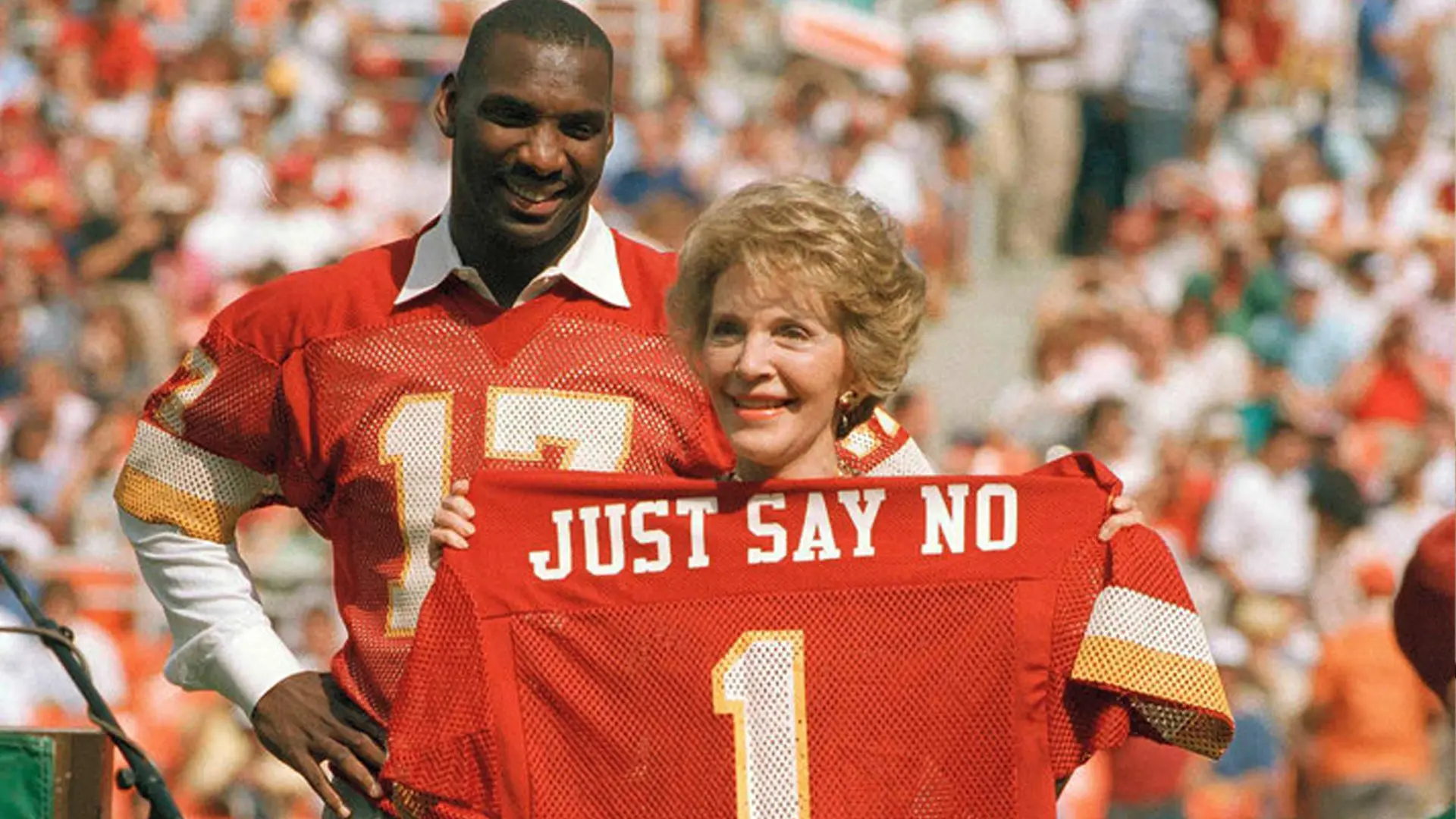 Two people in red sports jerseys hold up a jersey with "Just Say No" printed on it, standing on a sports field with a crowd in the background.