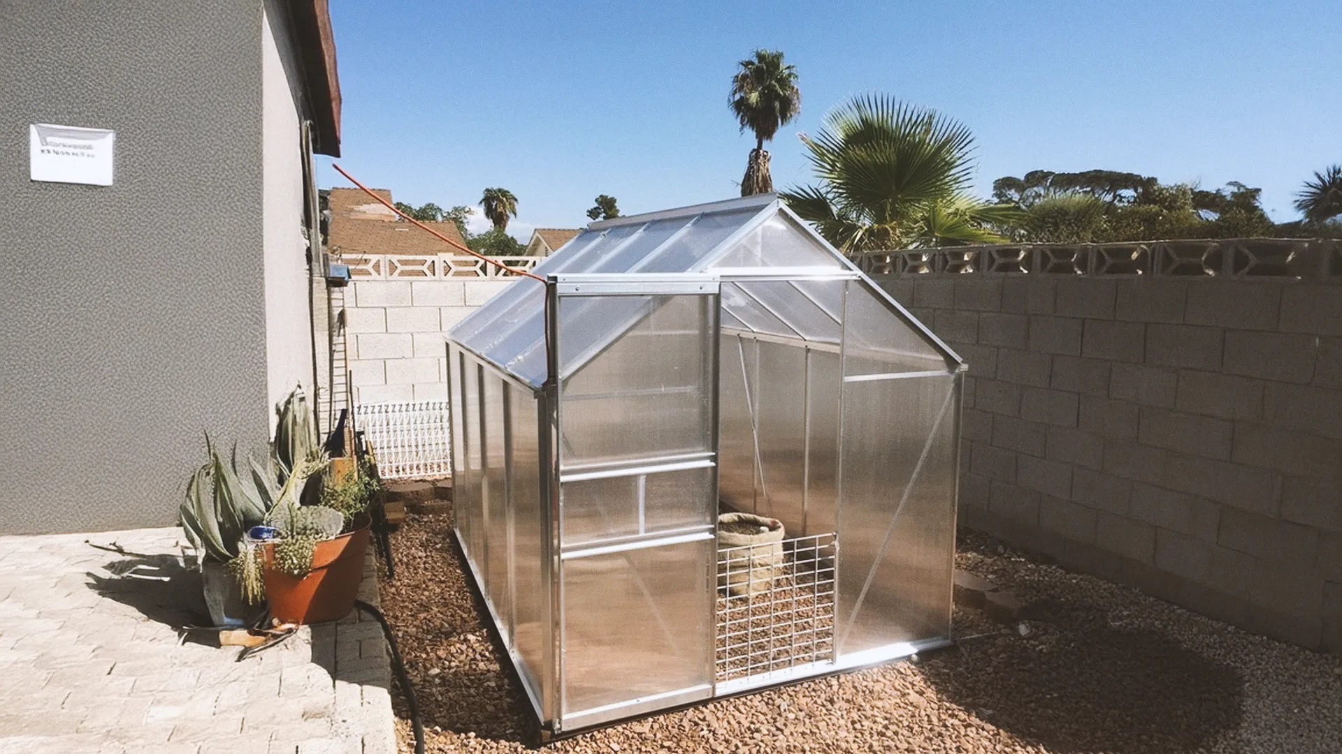 Small greenhouse with transparent panels in a backyard, surrounded by gravel and potted plants, under a clear blue sky.