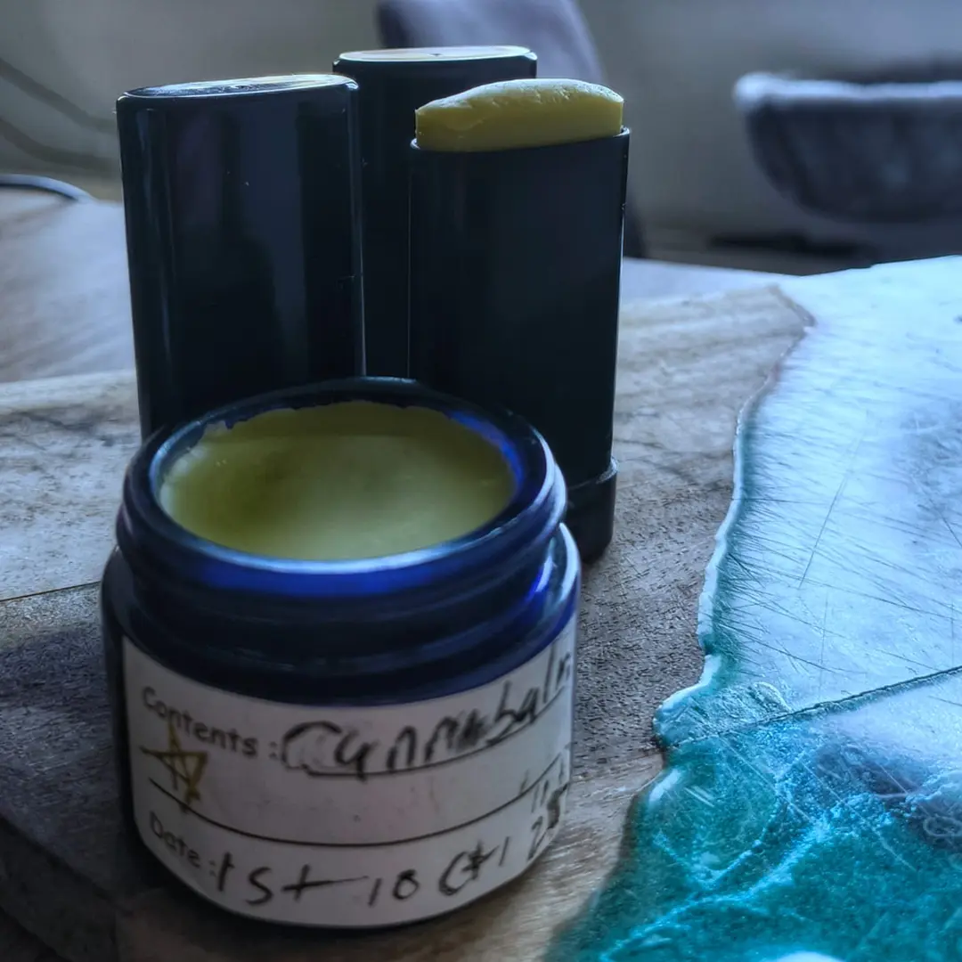 Open blue jar of yellow-green balm with handwritten label next to a black balm stick on a wooden table with a teal resin edge.